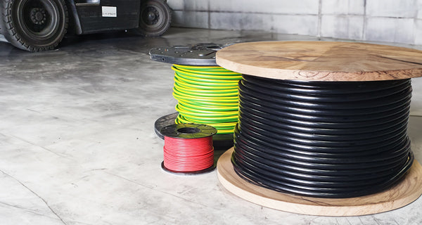 Black, earth, and red cable drums of various sizes in a warehouse with a forklift in the background.